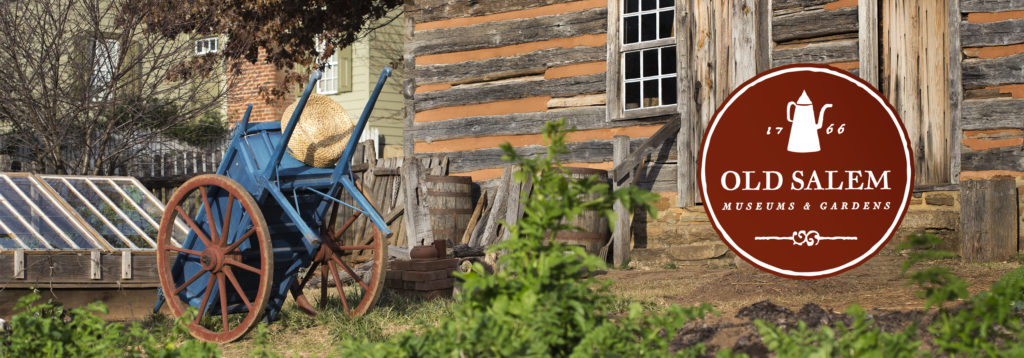 Blue wagon tipped over in front of log house. The Old Salem Museum & Gardens logo floats on the left side. 