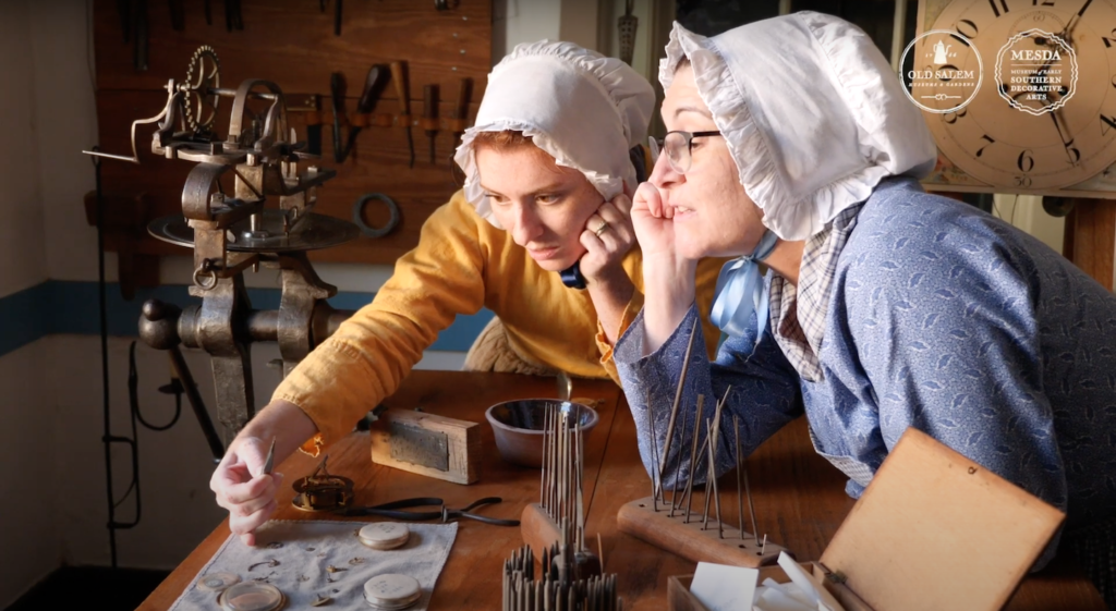 Two women in 18th century costume examine a historic object over a table with assorted tools. 