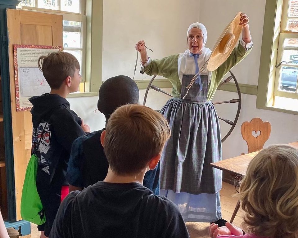 Woman in living history costume stands in front of a spinning wheel and holding a hat, speaking to students. 